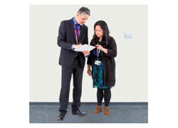 A man in a suit talking to a woman in a blue dress. They are reading a document.